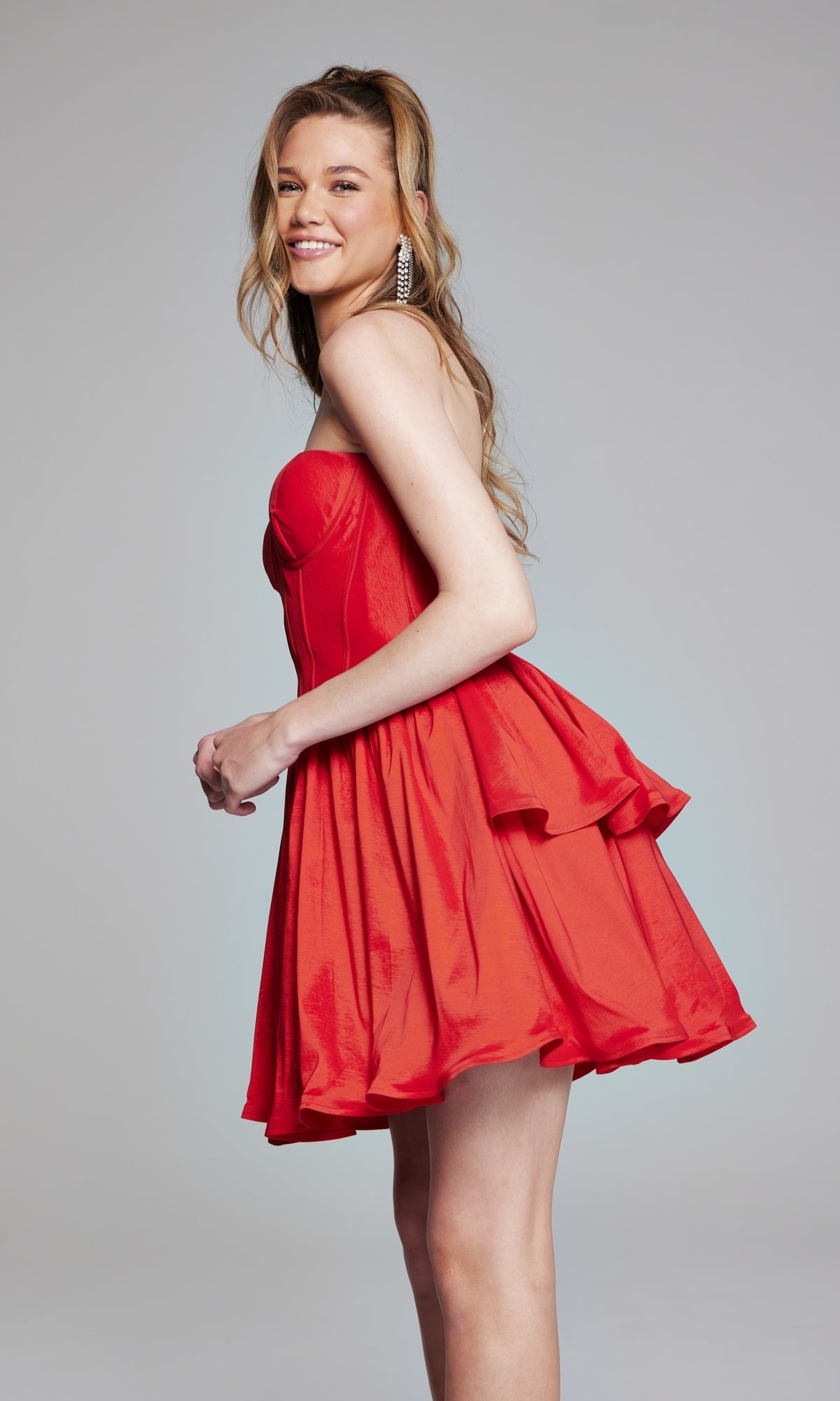 A woman smiles while standing sideways in front of a gray background, modeling the Jovani Strapless Short Ruffled Hoco Dress 36620, which features a strapless neckline and a playful ruffled skirt.